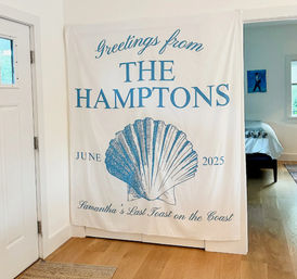 Coastal entryway banner reading 'Greetings from THE HAMPTONS' with a large blue scallop shell and 'June 2025', hanging over hardwood floors next to a doorway into a bedroom.