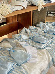 Blue-and-white striped satin pajama sets with light-blue satin drawstring pouches and white socks laid out on a wooden bench — beachy welcome-gift display.