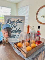 Austin, TX bachelorette party welcome display: framed playful welcome sign and oversized face cutout on a dining table beside a wooden tray holding champagne, Aperol, bottled water, mini liquor bottles, plastic champagne flutes and fresh oranges.