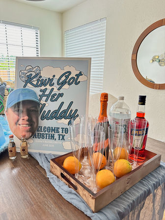 Austin, TX bachelorette party welcome display: framed playful welcome sign and oversized face cutout on a dining table beside a wooden tray holding champagne, Aperol, bottled water, mini liquor bottles, plastic champagne flutes and fresh oranges.