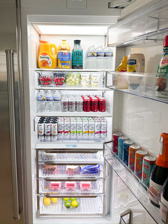 Open stainless-steel kitchen refrigerator with neatly arranged shelves of juices, cold brew, bottled water, sodas, canned seltzers, yogurt cups, berries and fresh fruit.