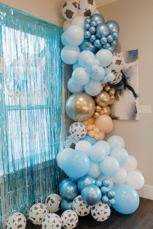 Indoor party backdrop in a home corner featuring a cascading organic balloon garland in shades of blue, white, gold and metallic teal with cow-print balloons beside a shimmering teal fringe curtain