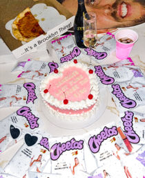 Two-tier pink bachelorette cake with white piped frosting and cherries on a table surrounded by themed snack bags, a champagne flute, a pizza slice in a box labeled "...It's a Brooklyn Thing!", heart-shaped sunglasses and a pink cup