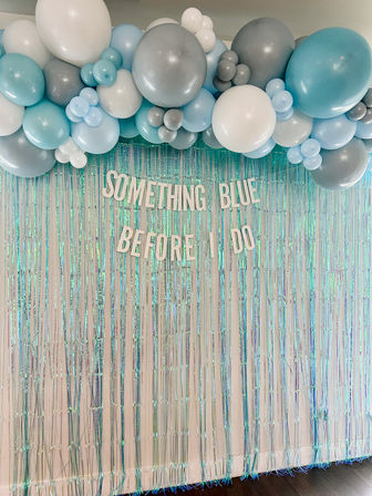 Playful indoor bridal shower photo wall: teal, baby blue, white and silver balloon garland over an iridescent fringe curtain with letters reading "Something Blue Before I Do".