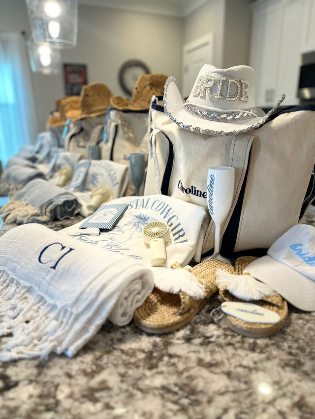 Coastal cowgirl bachelorette gift spread on a granite kitchen island: canvas tote bags topped with a white sequined “BRIDE” cowboy hat, personalized champagne flutes, beach towels, straw sandals and themed accessories.