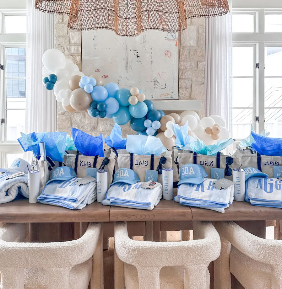 Bright coastal party display with a blue-and-beige balloon garland over a wooden table lined with personalized tote gift bags, 30A caps, monogrammed towels and tumblers in a sunlit beach-style dining room
