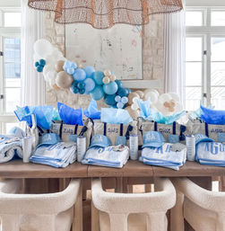 Bright coastal party display with a blue-and-beige balloon garland over a wooden table lined with personalized tote gift bags, 30A caps, monogrammed towels and tumblers in a sunlit beach-style dining room