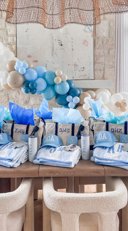 Bright coastal party display with a blue-and-beige balloon garland over a wooden table lined with personalized tote gift bags, 30A caps, monogrammed towels and tumblers in a sunlit beach-style dining room