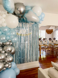 Cloud-themed baby shower decor in a home living room: blue, white and silver balloon arch, metallic tinsel backdrop and favor bags on a dining table.