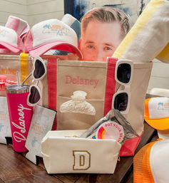 Beach-themed personalized canvas gift tote with pink trim packed with white sunglasses, a pink travel tumbler, a gold-initial pouch, trucker hats, striped beach towel, hair claw clip and party stickers — styled for a beach bachelorette or group getaway.