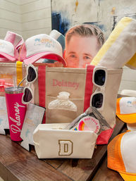 Coastal-themed summer gift bag on a wooden table: personalized canvas tote with pink trim and gold initial D, white heart and rectangular sunglasses, pink insulated tumbler, beach-logo trucker hats, rolled yellow towel, cosmetic pouch, shell hair clip, party stickers and a cardboard face cutout — bright pink, yellow and orange party favors.