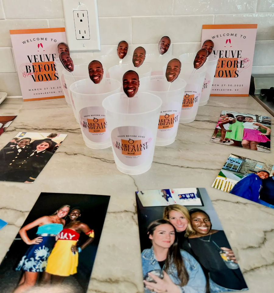 Plastic party cups with smiling face photo toppers arranged on a marble countertop, surrounded by printed photos and welcome event cards against a white subway tile backsplash.