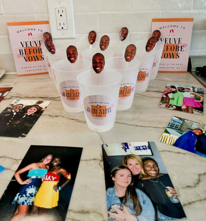 Plastic party cups with smiling face photo toppers arranged on a marble countertop, surrounded by printed photos and welcome event cards against a white subway tile backsplash.