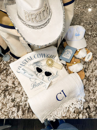 Coastal bachelorette flatlay featuring a white sequin "BRIDE" cowboy hat, canvas tote, coastal cowgirl tee, monogrammed fringe beach towel, white bride cap, straw tassel sandals, sunglasses, handheld fan and champagne glass arranged on a granite countertop — playful seaside bridal party packing.
