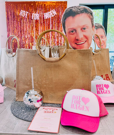 Event gift table with jute tote bags, disco-ball drink cups and bright pink trucker hats reading “Hot for Hagen,” oversized smiling face cutouts, and a pink foil fringe backdrop with matching slogan.