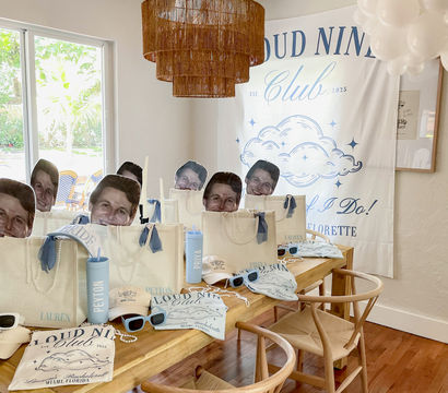 Boho-chic dining room setup for a Miami bachelorette party with a wooden table arranged with custom tote gift bags, oversized face cutouts on sticks, blue sunglasses and tumblers, cloud-themed banner and white balloons.