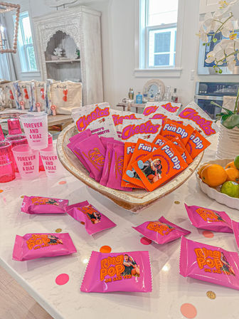 Bright kitchen island styled for an engagement party with a bowl of Cheetos and Fun Dip packets, scattered pink Ring Pop wrappers that say “She Said Yes!”, matching pink party cups on a marble countertop, fresh fruit bowl and white cabinets in the background.