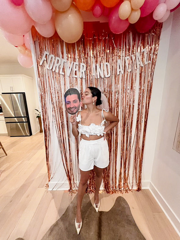 Bride-to-be in a white two-piece outfit and veil kisses a large cutout face on a stick in front of a rose-gold fringe backdrop with pink and peach balloons at an indoor bachelorette party.