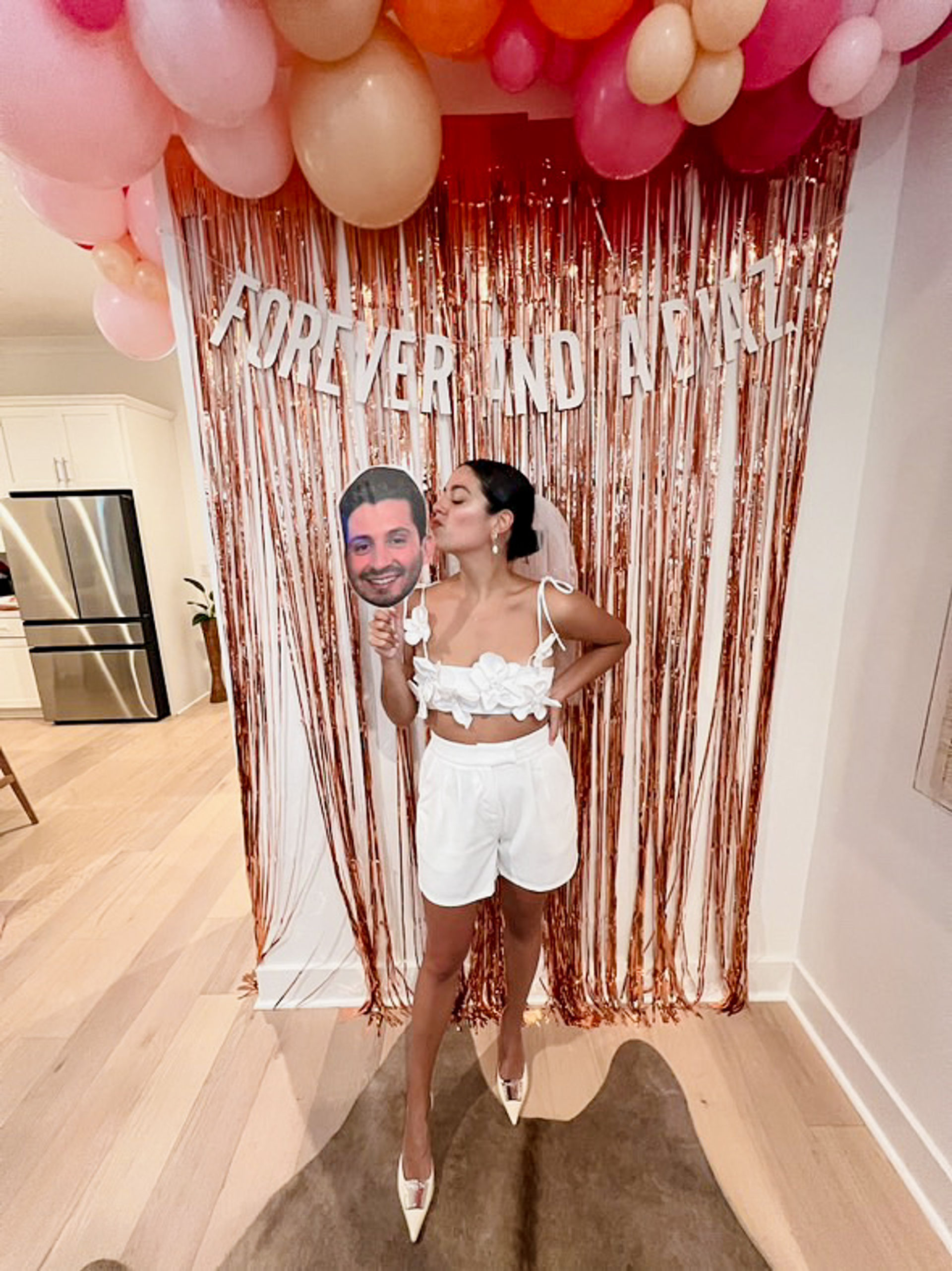 Person in a white bridal-style crop top, shorts, veil and heels kissing a large face cutout on a stick in front of a rose-gold fringe backdrop, pink and peach balloons and a banner reading "FOREVER AND ALWAYS" in a home setting