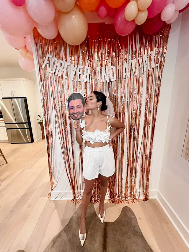 Person in a white bridal-style crop top, shorts, veil and heels kissing a large face cutout on a stick in front of a rose-gold fringe backdrop, pink and peach balloons and a banner reading "FOREVER AND ALWAYS" in a home setting