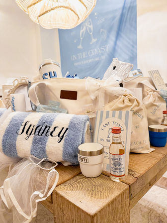 Coastal-themed bachelorette gift display on a rustic wooden table featuring a blue-and-white striped personalized towel, insulated cup, mini liquor bottle, canvas tote, drawstring favor bags and bridal veil