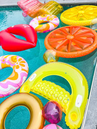Colorful inflatable pool floats — citrus slice, yellow lounge, gold ring, floral donut and red lips — floating in a turquoise backyard pool on a sunny summer day.