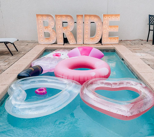 Backyard pool with large illuminated BRIDE marquee letters and pink-themed inflatables, including heart-shaped and donut-style floats for a bachelorette pool party.