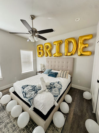 Bright modern bedroom decorated for a bride with gold foil balloons spelling BRIDE above a tufted bed, white balloons scattered on the floor, a towel swan and a blue patterned throw on white bedding.