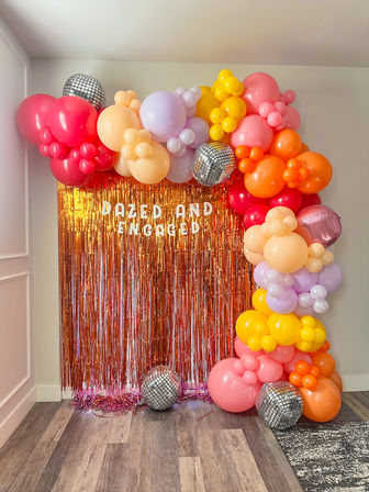 Indoor engagement party photo backdrop with colorful balloon garland in pink, orange, yellow and lavender, disco balls, and a rose-gold tinsel curtain reading 'DAZED AND ENGAGED'