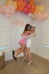 Two women hugging and laughing at an indoor bachelorette party photo booth under a pink, orange and yellow balloon arch with a white fringe backdrop; one wears a white cowgirl hat.