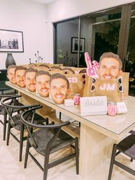 Playful bachelorette prep: row of personalized burlap gift bags and pink party favors on a long dining table, each topped with an oversized smiling male face cutout, sunglasses and a glittery 'Bride' pouch in a modern dining room.
