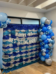 Blue and silver indoor bridal-shower photo backdrop with sequin fringe and white letters reading 'MAMMA MIA SHE'S GETTING MARRIED', accented by a cascading blue balloon garland and disco-ball decorations