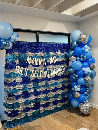Blue and silver indoor bridal-shower photo backdrop with sequin fringe and white letters reading 'MAMMA MIA SHE'S GETTING MARRIED', accented by a cascading blue balloon garland and disco-ball decorations