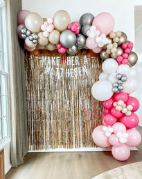 Festive indoor balloon arch in pink, blush, champagne and silver framing a gold fringe photo backdrop with a white letter banner, staged in a living room as a party photo booth on hardwood floor