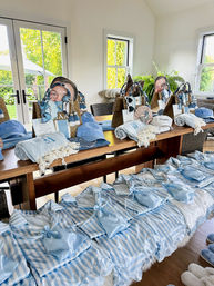 Sunlit dining room table arranged with coastal-themed welcome gifts: blue-and-white striped pajama sets, satin pouches, light-blue caps, woven beach totes, towels and slippers for a seaside weekend.