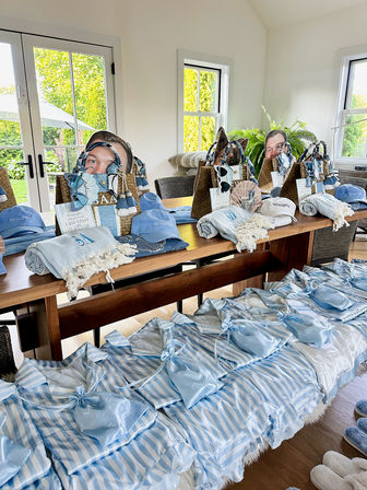 Sunlit dining room table arranged with coastal-themed welcome gifts: blue-and-white striped pajama sets, satin pouches, light-blue caps, woven beach totes, towels and slippers for a seaside weekend.