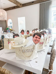 Row of personalized straw tote welcome bags filled with tissue and oversized smiling face cutouts on a white island in a bright coastal-style kitchen with exposed wooden beams and wicker chairs — beach getaway gift setup.