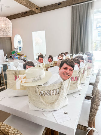 Row of personalized straw tote welcome bags filled with tissue and oversized smiling face cutouts on a white island in a bright coastal-style kitchen with exposed wooden beams and wicker chairs — beach getaway gift setup.