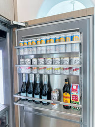 Open stainless steel kitchen refrigerator door revealing neatly arranged rows of canned hard seltzers, several sparkling wine bottles on the lower shelf, plus a bottle of coffee liqueur and a carton of half‑and‑half.