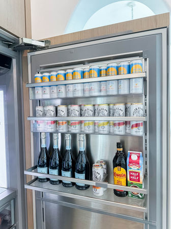 Open stainless-steel kitchen refrigerator door shelf neatly stocked with rows of canned seltzers (High Noon and White Claw), flavored seltzer cans, four sparkling wine bottles, a bottle of Kahlúa and a carton of half-and-half.