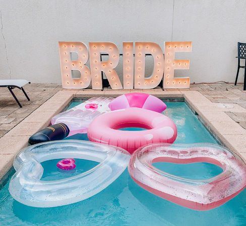 Backyard poolside bachelorette setup with pink donut and heart-shaped clear inflatables, a champagne bottle float, and large lit marquee letters spelling BRIDE on the patio.