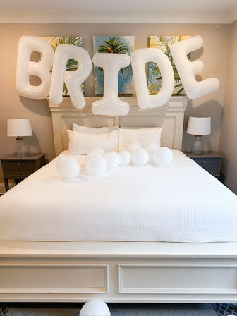 White foil letter balloons spelling 'BRIDE' float above a crisp white bed in a neutral bridal suite, with round white balloons on the mattress, bedside lamps and tropical wall art — bachelorette/wedding prep scene.