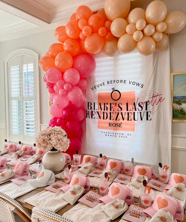 Rosé-themed bachelorette brunch setup in Charleston: pink and peach balloon arch over a decorated backdrop, long table lined with matching pink caps, tote bags, gift boxes, cups and a hydrangea centerpiece.
