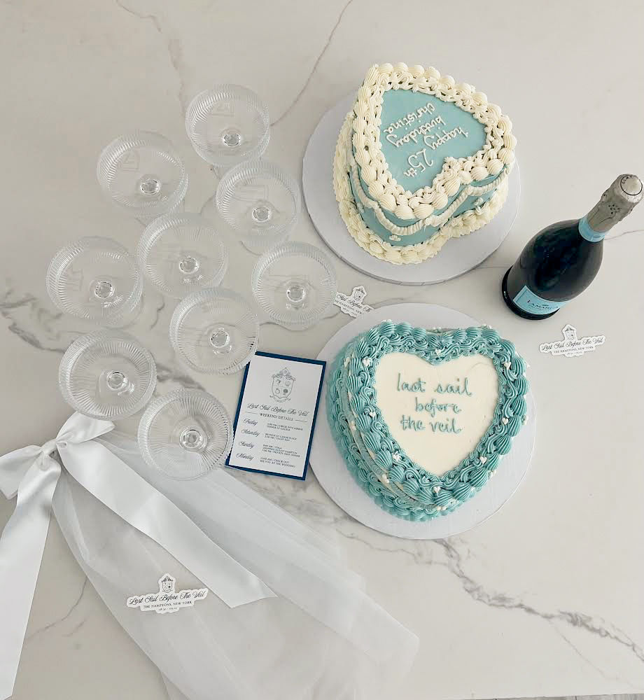 Overhead shot of a bridal shower table on marble with two blue-and-white heart-shaped cakes (one reads last sail before the veil), a bottle of champagne, stacked coupe glasses, white veil ribbon and an invitation card