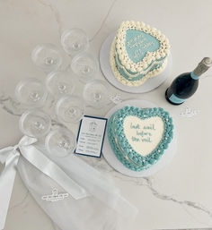 Overhead shot of a bridal shower table on marble with two blue-and-white heart-shaped cakes (one reads last sail before the veil), a bottle of champagne, stacked coupe glasses, white veil ribbon and an invitation card