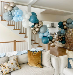 Open-concept living room in a modern home with teal, light-blue, white and metallic gold balloon garland draped along a wooden staircase and doorway above a neutral sofa with velvet and patterned throw pillows.