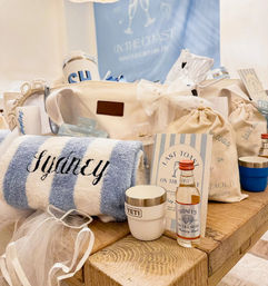 Coastal bachelorette welcome kit on a rustic wooden table with blue-and-white striped spa towel, insulated travel cup, mini liquor bottle, drawstring favor bags, striped “Last Toast on the Coast” card and a canvas tote in soft beach colors.