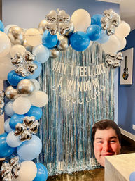 Indoor party photo backdrop with blue, white and metallic silver balloons and star-shaped foil accents forming an arch around a shimmery blue fringe curtain, white letter banner, and a large smiling face cutout