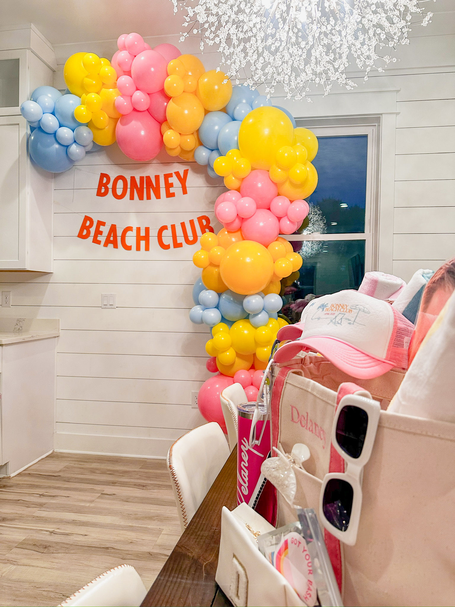 Indoor coastal-style celebration with pastel pink, yellow and blue balloon arch framing a 'Beach Club' banner, chandelier overhead and a table with pink hats, sunglasses and gift totes.