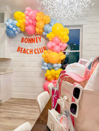 Indoor coastal-style celebration with pastel pink, yellow and blue balloon arch framing a 'Beach Club' banner, chandelier overhead and a table with pink hats, sunglasses and gift totes.